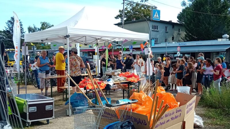 Menschen stehen unter einem weißen Paviliion. Im Vordergrund sind Gerätschaften zum Müll Sammeln sichtbar. Rechts von dem Pavillion steht eine Gruppe von Menschen.