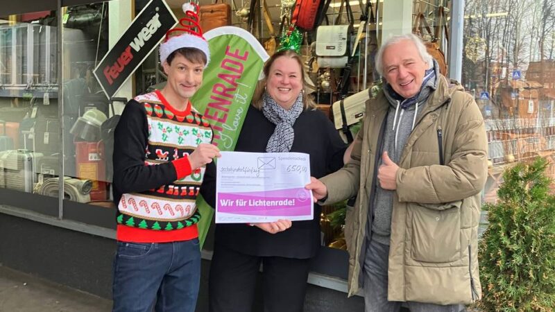 Drei Menschen halten die Spendenbescheinigung in den Händen, die 2025 an den Blohmgarten Lichtenrade ging. Im Hintergrund des Bildes ist ein Geschäft sichtbar, in dem Taschen und andere Artikel verkauft werden.